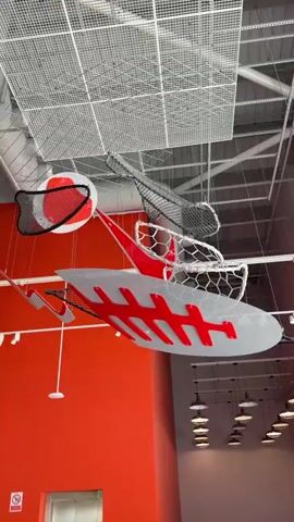 The video pans across a wide indoor space, likely a gymnasium or recreational facility, focusing on a large, abstract art installation suspended from the ceiling. The installation features several net-like structures and large, flat, brightly colored components, predominantly red and white, with grey accents. One prominent element resembles a stylized shark or fish skeleton, with a large, grey, fin-like body adorned with bold red rib-like markings. Another section appears to be a suspended seating area or lounge, with red and white curved structures that look like chairs or pods, also outlined in netting. A red and white sphere, partially enclosed by black netting, is also visible. The ceiling is a grid of white metal mesh, revealing industrial lighting fixtures and air ducts. The walls are painted in a vibrant orange on the left side of the frame, transitioning to a neutral grey on the right. A series of dark pendant lights hang in rows along the right wall, illuminating the space. The camera movement is smooth, a slow pan and slight tilt, allowing the viewer to take in the scale and detail of the installation. No people are visible in the video.