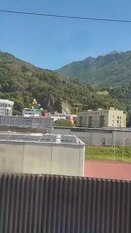 The video captures a view from a moving vehicle, likely a train, showing an urban environment with buildings and a backdrop of lush green mountains under a clear blue sky. Several colorful balloons are visible, floating above some of the buildings. In the foreground, there is a striped barrier or fence. The scene suggests a daytime view during fair weather. There are no identifiable people or direct interactions depicted.