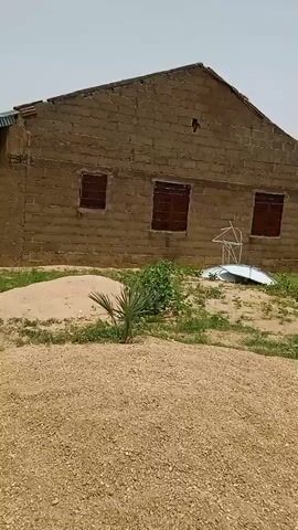 The video shows the exterior of a one-story building constructed of tan brick. The building has a peaked roof with a corrugated metal overhang. Several dark brown framed windows are visible. The building sits on a ground of gravel and sparse green vegetation. A small pile of sand and a small palm tree are in front of the building. A metal frame structure lies on the ground next to the palm tree. In the background, there is a flat, barren field and other buildings are visible in the distance. The sky is light gray. The camera pans slowly to the right.