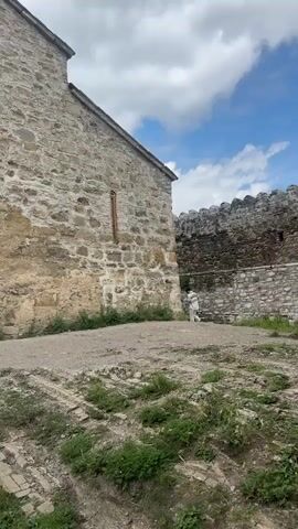 The video shows a stone building with arched windows and a conical roof. The building appears to be old and is made of rough stone. There are two people walking on a path in front of the building. One person is a woman wearing a black t-shirt and shorts. The other person is a man wearing a black t-shirt and shorts. They are walking away from the camera. There is a wall in the background made of stone. The sky is cloudy and there are some trees in the background.