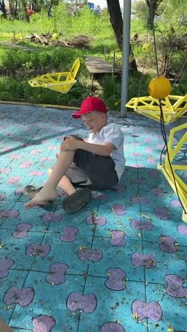 A boy is sitting on a playground floor made of blue tiles with purple flower patterns. He's wearing a red baseball cap, a white t-shirt, and dark gray shorts. He smiles and appears to be grabbing his leg. He has one shoe on and another shoe beside him. There are two yellow swings in the background. The area is outdoors, with green grass and trees visible. The boy stands up with the help of his hands and feet on the ground.