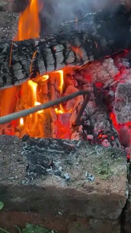 A close-up shot of a campfire shows logs burning intensely, with flames reaching upwards. Embers glow red, and sparks fly from the fire. A metal rod is inserted into the burning logs, suggesting someone is tending to the fire or cooking. The fire is contained within a structure made of bricks. The surrounding area appears to be outdoors, with some grass visible at the bottom of the frame. The smoke from the fire drifts upwards and to the right. The lighting suggests it is either daytime or a well-lit evening.
