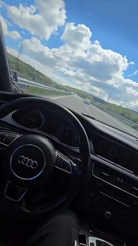 The video shows the perspective of someone driving an Audi on a highway during daytime. The sky is blue with fluffy white clouds. The road is clear with a few other vehicles visible in the distance. Green hills line the sides of the highway. The car's dashboard and steering wheel are in focus. The steering wheel has the Audi logo in the center. Music is audible in the background. The driver's leg is visible in the bottom left corner of the frame.