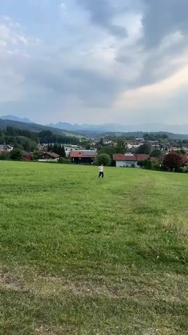 A young boy is running through a grassy field. He is wearing a white t-shirt and dark pants. The boy bends down, then stands up, holding a stick. He continues to run and play in the field. In the background, there is a village with houses and trees, and mountains in the distance. The sky is overcast.