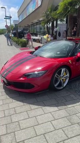 The video shows a red Ferrari 296 GTS parked in front of a building with "SUMMER VIBES" in the window. The car has black racing stripes and yellow brake calipers. The building has a sign for ITALIAN TANALYSE. There are palm trees and a few people in the background. One person is taking a picture of the car. There are balloons near the entrance. The ground is paved with gray bricks.