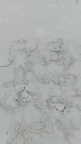 The video shows an overhead view of a sandy surface with the words "RED JOINS COLLEGE MEME" written in the sand. A brown sandal appears at the bottom of the frame. The sandy surface is textured and uneven. The camera remains mostly static throughout the video. There is a light-colored grainy texture on the sandy surface.