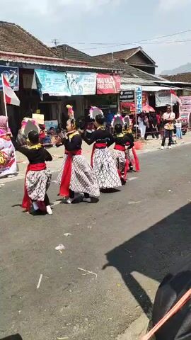 A group of young girls in traditional Indonesian attire are performing a dance in the street. They are wearing colorful headpieces, sarongs, and red sashes. The girls are moving in unison, their arms outstretched and their bodies in graceful poses. The street is lined with spectators, and buildings can be seen in the background. The weather appears to be sunny.