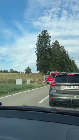 The video shows a view from inside a car looking out at a rural landscape. A red train is passing by on tracks to the left of the road. The train has several windows and doors, and text that appears to be in French. The car is stopped, indicated by the traffic and the presence of other vehicles. The sky is blue with some clouds. There are trees and fields visible in the background. The video captures the moment the train passes the stationary car.