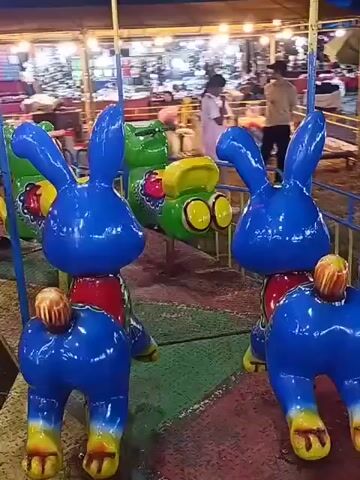 This video shows a carousel ride at what appears to be a fair or amusement park. The carousel features several brightly colored, cartoon-like figures. In the foreground, there are two blue rabbit figures, each with large ears, a red collar, and a yellow tail. They are standing on four legs, with their backs facing the camera. Behind the rabbits, there are green frog-like figures and yellow horse figures. The carousel is illuminated by overhead lights, creating a vibrant atmosphere. Some people are visible in the background, appearing to be enjoying the rides or browsing the stalls. The ground beneath the carousel appears to be covered in a patterned mat or carpet. The overall impression is one of a lively and cheerful amusement setting.