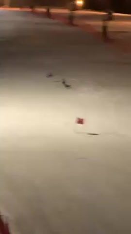 The video shows a skier moving downhill through a slalom course. The skier wears a dark-colored outfit and skis in a snow-covered environment with several red and blue poles marking the course. The illumination is poor, and the lighting is artificial. The skier makes turns, passing the poles.