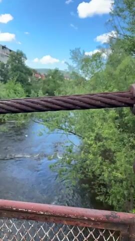 The video shows a river flowing under a bridge on a sunny day. The camera is positioned on the bridge, looking down at the water. The river is surrounded by green trees and bushes. In the background, there is a building and a hill. The sky is blue with some white clouds. The bridge's support cable and railing are visible in the foreground.