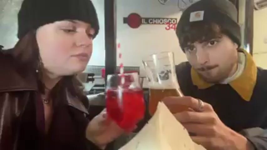 Two people, a woman and a man, are sitting at a table inside a cafe or restaurant. The woman is on the left wearing a black beanie, a brown coat, and earrings. She holds a wine glass filled with a red drink and a striped straw. The man is on the right, also wearing a black beanie, and a jacket with a yellow collar. He holds a beer glass. They are making a toast. Behind them is a sign that reads "IL CHIOSCO 34." The lighting is soft and natural.