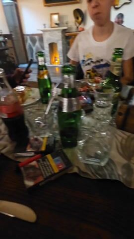 The video shows a table in what appears to be a restaurant, where several people are seated. On the table, there is a bottle of "Baldaran" water, various glasses with different liquids, a plate with a dish (possibly pizza), cutlery, napkins, and cell phones. Some of the people at the table are looking at their phones. The environment seems to be warm and relaxed, suggesting a casual dining setting. The restaurant has a dark wooden ceiling and indoor lighting.