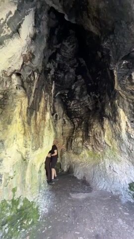The video begins inside what appears to be a hollow tree trunk, with layered rock-like formations on the inside. A person stands inside the hollow space wearing a black t-shirt and shorts. The video then transitions to an outdoor scene. People are walking along a dirt path near a body of water, possibly a lake or wide river. There are trees lining the path. One person is walking with a small dog. Another person stands near the water's edge, facing away from the camera. The overall environment suggests a park or natural area.