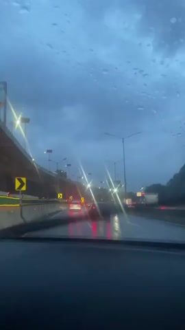 The video shows a view from the dashboard of a car driving on a multi-lane highway in Cuautitlán Izcalli, Mexico, during what appears to be dusk or nighttime. The sky is dark and overcast, suggesting rainy weather, which is confirmed by the wet windshield with visible raindrops and the glistening road surface. Traffic is moderately heavy, with several cars visible ahead, their red taillights glowing. Streetlights illuminate the highway, creating bright streaks of light due to the wet windshield.

To the left, an overpass is visible with more streetlights, and yellow chevron signs pointing to the right are mounted along the road's edge. The overall scene depicts a typical highway commute in challenging weather conditions, with the artificial lights reflecting off the wet surfaces.