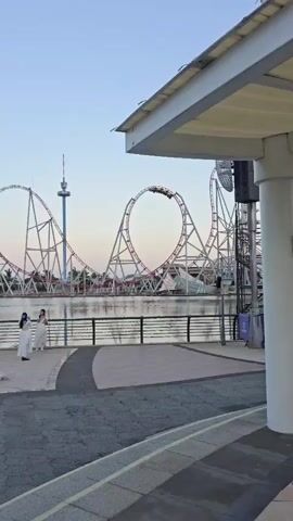 Two people in white dresses stand near a body of water. A roller coaster with several loops and a tall tower are visible in the background. The sky is a clear blue.