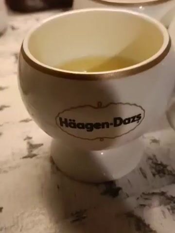 The video shows five white Haagen-Dazs ramekins filled with a yellow liquid. The ramekins are arranged in a semi-circle on a patterned surface. A red pot is visible in the background.