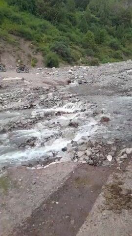 The video shows a rocky riverbed with a stream of water flowing through it. On the bank of the river, there is a dirt road. Several motorcycles are parked on the road, and one person can be seen riding a motorcycle. The surrounding environment is hilly and covered with green vegetation and trees. The sky is cloudy.