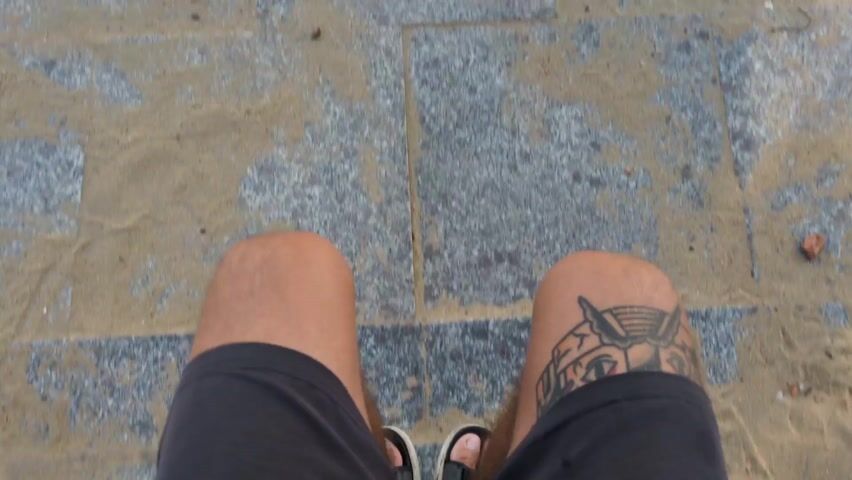 A first-person, downward-angle shot shows a person standing on a walkway composed of gray stone or concrete squares, which are partially covered in sand. The person is wearing black shorts and black sandals with white trim. A tattoo is visible on the right leg, just above the knee. The sand appears to be encroaching onto the walkway from the sides. The camera is static, focused on the legs and feet, capturing the texture of the ground and the tattoo detail.