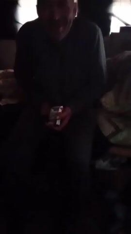 A dimly lit video shows a man sitting down. He is fair-skinned, has thinning hair, and a short beard. He is wearing a dark-colored, long-sleeved shirt. He holds a small red and white container in his hands. The man appears to be inside a poorly lit space with low ceilings and miscellaneous items scattered around, including what appears to be bedding. There's movement in the frame, indicating possible camera shake.