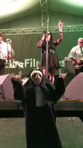 On a stage illuminated by bright white and green lighting, a nun figure stands in the foreground, making the "rock on" hand gesture. Behind the nun, a band performs, including a female vocalist, a guitarist, and a horn player. Large speakers flank the nun figure, and a white banner hangs in the background. The nun figure is wearing a black habit with a white wimple and a silver cross. The band members are dressed in a mix of casual and formal attire.