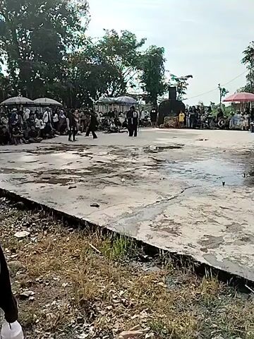 The video shows three individuals performing some sort of martial arts display on a concrete platform. The individuals are wearing black clothing and appear to be using some type of rope or whip as part of their performance. An audience surrounds the platform. The area is outdoors, with trees in the background, and food vendors on either side of the viewing area. The ground is wet, and there is debris scattered on the platform.