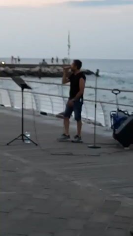A man in a black sleeveless shirt and denim shorts sings into a microphone on a boardwalk. A music stand is to his left. Another man in a black sleeveless shirt and jeans walks away from the camera towards the singer. A woman in a black tank top and white skirt walks past the camera. The ocean is visible in the background, along with a pier and other people. The sky is overcast.