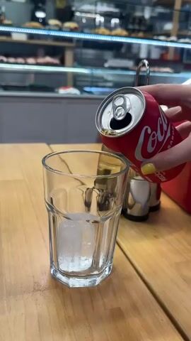 A person's hand, with yellow nail polish, holds a red Coca-Cola can and pours the beverage into a clear glass filled with ice. The glass is on a wooden table. In the background, there is a display case filled with food items and a red object, possibly a napkin dispenser. The pouring action creates some foam in the glass.