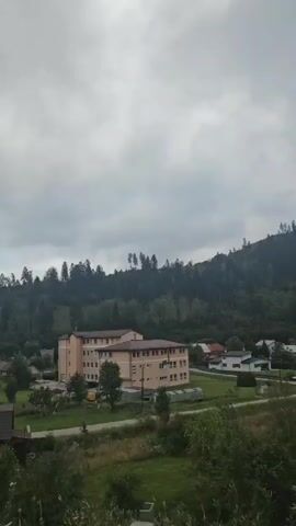 The video shows a landscape view with a building in the center, a wooded hill in the background, and some houses and vegetation in the foreground. The sky is cloudy. The building appears to be a multi-story structure, possibly a school or hospital, with a light-colored facade and a brown roof. There are trees and greenery surrounding the building and on the hill. A wooden fence is visible in the lower part of the frame. The time stamp indicates the video is very short, around 4 seconds.