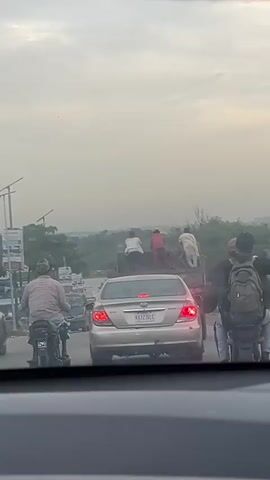 A video taken from inside a vehicle shows a traffic scene. A silver sedan is in the foreground, facing away from the camera. To the left of the sedan, two motorcycles are visible, with riders wearing casual clothing. To the right of the sedan, another motorcycle is partially visible, with a rider wearing a backpack. In front of the sedan, a flatbed truck is moving, carrying several people on its bed. The people are standing and appear to be dressed in light-colored clothing. The background shows a hazy sky and some trees. The overall impression is a congested road in what appears to be a developing country, possibly during the daytime.