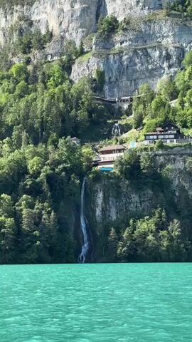 A drone shot moves past a turquoise lake towards a steep, forested cliff face. Perched on the cliff are several buildings with dark roofs, connected by winding paths and staircases. A thin waterfall cascades down the rock face, emptying into the lake. The mountain peak above is jagged and gray.