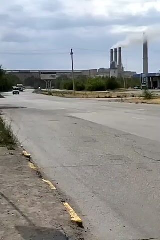 The video shows an overcast day with a large industrial complex in the background emitting smoke from several smokestacks. A paved road runs through the foreground with a dirt shoulder. Two vehicles are visible on the road; a white SUV is in the left lane, and a truck is in the right lane. Both vehicles appear to be moving away from the camera. The industrial complex consists of a long, low building and several tall smokestacks. The sky is filled with heavy clouds.