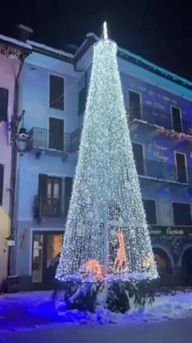 Here's a description of the video:
The video shows a large, illuminated Christmas tree made of lights, standing outdoors in a snowy town square at night. The tree is cone-shaped, predominantly white with some other colored lights and has a tall pole in the center. At the base of the tree, there are illuminated figures of deer and other animals. There is snow on the ground. Behind the tree is a building with multiple stories and windows. The building is painted in light colors and has decorative lights around the windows. There are some other buildings visible on the side. It appears to be snowing lightly. The scene is lit up by the Christmas tree and various other lights around the square.