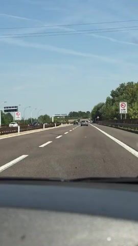 The video shows a first-person view of a car driving on a highway. The road is asphalt with white dashed lines indicating lanes. There are other cars on the road ahead, some of which are also moving. On the right side of the road, there are trees and a guardrail. On the left side, there is a guardrail and some signs. The sky is clear and blue with some clouds. The sun is shining, indicating it is daytime. The video is shaky, suggesting movement and possibly a passenger recording.