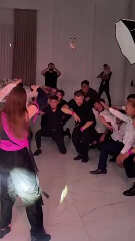 The video shows a group of people dancing and celebrating in what appears to be a formal event space. The room has white marble floors and is decorated with draped curtains and lighting. In the foreground, a young woman with long brown hair, wearing a pink top and black pants, is dancing. Several men in black clothing are gathered around her, some on their knees, reaching out towards her. One man in the center, also in black, is performing a move with his arms outstretched. A large softbox light is visible in the upper right corner of the frame. The lighting changes throughout the video, shifting from pink and white to blue and then back to pink. Other people are visible in the background, some dancing and some observing. In one segment, a line of women, some in dresses and some in casual wear, are shown dancing with their arms raised.