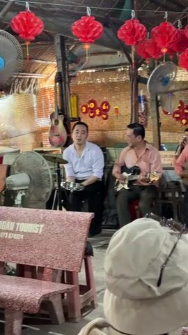 The video shows an open-air or semi-outdoor restaurant or cafe in Thới Sơn, Vietnam. Several red paper lanterns are strung across the ceiling, along with fans. A group of three men is performing traditional music; one plays a guitar, another a string instrument resembling a đàn nguyệt or similar Vietnamese lute, and a third man plays a small percussion instrument like a shaker or tambourine.

Guests are seated at stone-like tables and benches that have "CONG DOAN TOURIST" and a phone number ("T.073 874324") inscribed on them. On the tables are small plates of fruit (including watermelon, pineapple, and dragon fruit), teacups, and teapots, suggesting refreshments are being served.

Among the patrons, a woman in a light green shirt smiles at the camera. Two young boys are also visible at a table; one wears a white baseball cap and a red shirt, while the other, also in a red shirt, has some markings on his face and is holding a small cup. The overall atmosphere appears relaxed and festive, with live music entertaining the guests.