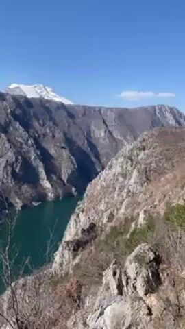 The camera pans down from a mountain peak with snow to a turquoise body of water, potentially a river or a lake, nestled between rocky cliffs. The landscape is rugged, with sparse vegetation clinging to the steep slopes. The sky is clear and blue. The scene evokes a sense of natural beauty and remote wilderness.