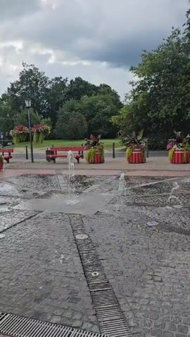 A public square features a splash pad with multiple jets of water spraying upward from the cobblestone ground. In the background, a row of red benches is partially visible, interspersed with large red planters filled with greenery and purple-red flowers. Trees and a cloudy sky form the backdrop. The scene is captured from a low angle, focusing on the water features.