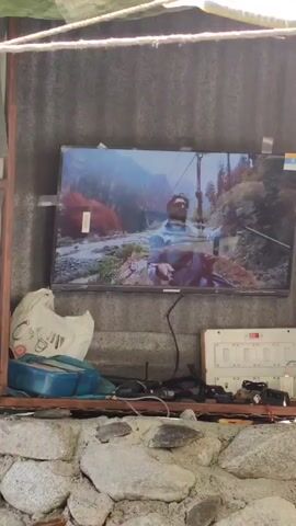 The video shows an outdoor scene. In the first frame, a television is mounted on a corrugated metal wall, displaying a man in a blue shirt against a mountain landscape. Below the TV are various objects, including a plastic bag, a blue container, cables, and a power strip. The second frame shows a broader view of the area with a small structure and people sitting on chairs. The third frame focuses on a sign that reads "HIGHEST & LONGEST ZIPLINE" with stairs leading up to it. The fourth frame shows a view of trees, a sky, and mountains in the distance, with a bamboo pole in the foreground.