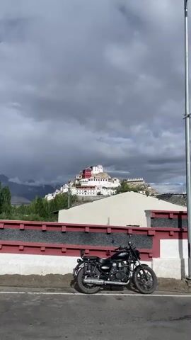 A black Royal Enfield motorcycle is parked on the side of a paved road, facing right. Behind the motorcycle is a low wall with a red and white pattern. Further in the background, a white and red building complex is situated on a hilltop. To the right of the motorcycle, a tall metal pole stands vertically. The sky is cloudy with dark storm clouds gathering. The scene transitions to show a road winding up a rocky, barren hillside with scattered green trees and a few distant figures walking up the road.