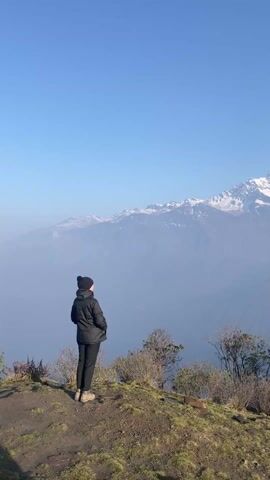 A person stands on a hilltop overlooking a vista of snow-capped mountains on a clear, sunny day. The person, seen from the back, wears a black jacket, pants, boots, and a beanie. They are positioned on a patch of grassy land dotted with small shrubs and plants. In front of them, a valley stretches out, partially obscured by a light haze. The background is dominated by a large mountain range covered in snow and rock, clearly visible against the clear blue sky.