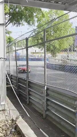 A red and white race car is shown on a race track, viewed through a chain-link fence. The car is moving and appears to be in motion. The track has guardrails on the side. Trees and overhead structures are visible in the background. The scene appears to be outdoors during the daytime.