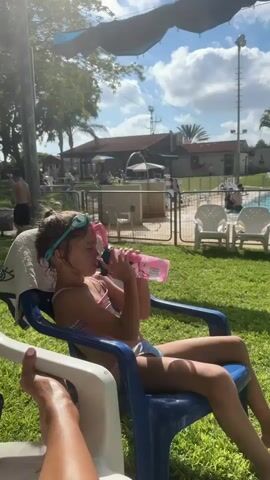 A young girl in a pink bikini and turquoise goggles sits in a blue plastic chair, drinking from a pink water bottle. She is outdoors, seated on a grassy lawn with a white towel draped over the back of her chair. In the background, a swimming pool is visible, with lounge chairs and people around it. A fence separates the pool area from the grassy area where the girl is sitting. Trees and buildings are also visible in the distance. The sun is shining, casting shadows and suggesting it is a bright, warm day. The sky is partly cloudy. The overall mood appears relaxed and summery, likely a day at a pool or water park. There is no visible text in the image.