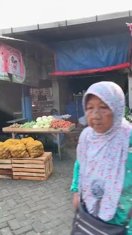 The video shows a bustling outdoor market scene. In the foreground, a person wearing a light-colored hijab and a patterned jacket walks away from the camera. They are also wearing a yellow garment underneath and a black skirt. To their left, a stall displays a variety of fresh produce, including carrots, leafy greens, and cabbage, arranged in crates. In the background, more market stalls are visible, with awnings providing shade. Various goods, including baskets of produce, bags of grains, and red woven containers, are displayed. A black motorcycle is parked in the lower left corner of the frame. The market appears to be active, with multiple people in the background, although their features are not clearly discernible. The overall environment suggests a typical day at a local market.