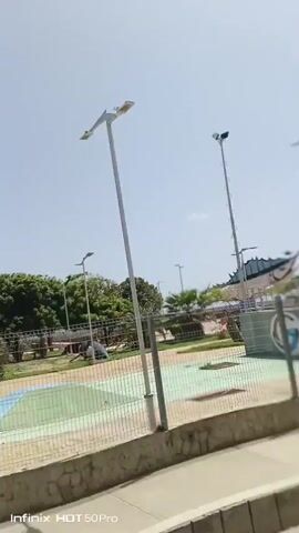 This video captures a bright, sunny day in Macuto, Venezuela. The scene appears to be an outdoor recreational area or park, possibly near the coast, given the expansive sky and distant view of what could be water. 

Several tall lampposts are prominent features throughout the video, suggesting the area is equipped for evening use. In the foreground, a wire mesh fence runs along a low concrete wall. Beyond the fence, there are paved areas marked with colorful sections, resembling a court for sports or games. 

Scattered throughout the park are various trees, including palm trees, and some smaller shrubs and grassy patches. In some frames, there are covered seating areas or gazebos with red roofs. There is also a tall structure with a ladder, possibly a lookout tower or a specialized lighting tower.

The overall atmosphere is calm and open, with no people visible in the footage. The clear blue sky indicates fair weather. The text "Infinix HOT 50Pro" is visible in the bottom left corner of each frame, indicating the device used to capture the video.