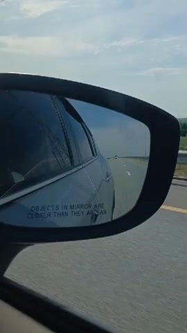 The video captures a first-person perspective from inside a moving vehicle, focusing on the side-view mirror. The mirror reflects a silver sedan driving alongside, with the phrase "OBJECTS IN MIRROR ARE CLOSER THAN THEY APPEAR" visible. Beyond the car in the mirror, a highway stretches into the distance under a partly cloudy sky. The right side of the frame shows the actual view from the car, which is also a highway with a grassy median and a guardrail. The landscape outside consists of rolling green hills and scattered trees. The overall impression is one of travel on a sunny day.