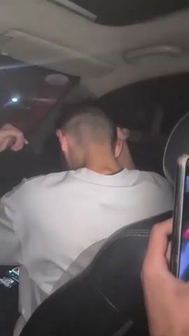 A man is visible in the back seat of a car, facing away from the camera. He wears a light-colored t-shirt, has short dark hair, and appears to be dancing or moving his arms rhythmically. Another person, presumably holding a smartphone, is to the side of the man and briefly shines the phone's flashlight on him. The car has a sunroof, and through the back window, taillights of another car can be seen. The interior of the car is dark. The overall impression is a candid, possibly celebratory moment in a moving vehicle.