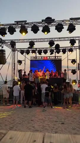 The video opens with a close-up of a stage with multiple spotlights overhead. A DJ booth is visible on the stage, with several people gathered around it. The crowd is on a sandy beach, with the ocean in the background. As the video pans to the right, more of the beach is revealed, with some barrels and lighting equipment. The sky is a soft gradient of orange and pink, indicating sunset. People are dancing and socializing in front of the stage. The overall atmosphere is that of an outdoor beach party or concert.