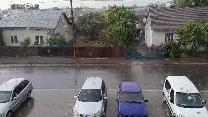 This media file shows an outdoor scene during a rain shower. The foreground displays several parked cars: a silver hatchback, a blue sedan, a white van, and a dark-colored sedan further to the right. A white SUV is seen driving on a wet road in the middle ground. Behind the road, a wooden fence separates the street from residential properties. Several houses with tiled roofs are visible, surrounded by lush green trees. The weather is clearly rainy, with visible raindrops on the lens and wet surfaces, suggesting it is daytime. The scene appears to be a street in a residential area, possibly a town or village, given the houses and parked vehicles. There are no people visible in the frame, and the overall mood is tranquil despite the rain, focusing on the quiet street activity. No visible text is present in the video. The location is identified as Pustomyty, Ukraine.