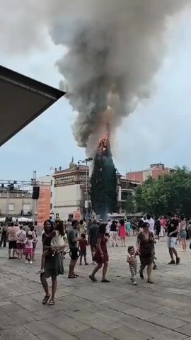 The video shows a tall cypress tree engulfed in flames, emitting a large column of dark smoke into a cloudy sky. The tree is situated in an outdoor plaza paved with cobblestones, with buildings in the background. A crowd of people, including adults and children, are gathered in the plaza, many observing the fire. Some appear to be holding phones up, possibly recording the event. The atmosphere seems to be one of shock and curiosity, with people standing at a distance from the burning tree. A stage with a banner is visible to the left, suggesting a public event or festival. The presence of a bicycle and people in casual summer clothing indicates it is likely daytime in warm weather. The setting appears to be a town square in Sant Cugat del Vallès, Spain, given the provided location context. No text is clearly legible on the banner. The event is clearly a fire in a public space, with a large tree as the focal point.
